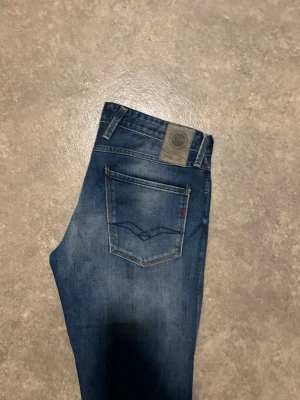 Replay Anbass blå jeans, W34 L32 - Säljer ett par Replay Anbass jeans i klassisk blå tvätt med snygga slitningar. Modellen har fem fickor, normal passform och raka ben. Materialet är mjukt jeans-tyg i bomull och polyester. Perfekta för dig som vill ha ett par stilrena och bekväma jeans.