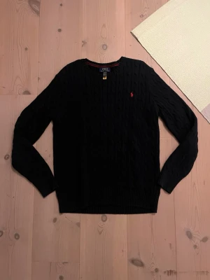 Svart kabelstickad tröja Polo Ralph Lauren - Kabelstickad svart tröja från Polo Ralph Lauren i storlek XL. Klassisk rund halsringning, långärmad och med röd broderad logga på bröstet. Tillverkad i mjuk bomull som känns skön mot huden. Perfekt för dig som gillar stilrena och tidlösa plagg.