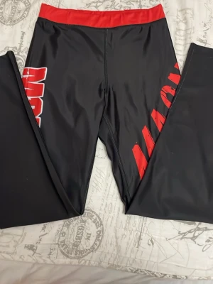 Svarta leggings med röd midja och tryck - Säljer ett par svarta leggings med bred röd midja och stort rött tryck längs benen. Materialet är stretchigt och passar perfekt för träning eller chill. Snygg kontrast mellan svart och rött gör dem riktigt iögonfallande.