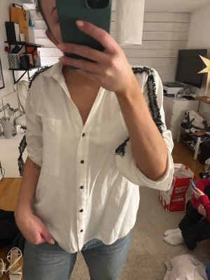 Vit skjorta med fransdetaljer Zara - Vit skjorta från Zara Basic i bomull med svarta fransdetaljer längs axlar och ärmar. Klassisk krage, svarta knappar framtill och en bröstficka. Skjortan har lång ärm och en loose passform som ger en chill vibe.