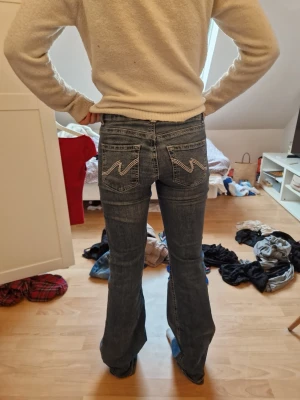 Blå bootcut jeans med broderade fickor - Säljer ett par blå bootcut jeans med låg midja och snygga broderade detaljer på bakfickorna. Jeansen har dubbla knappar framtill och klassisk femficksdesign. Perfekta för dig som gillar en avslappnad men ändå trendig look.