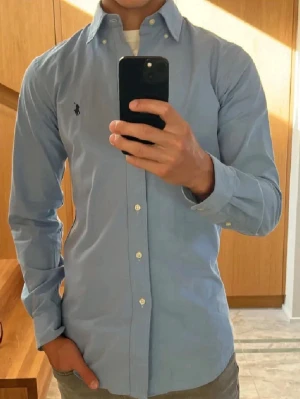 Polo Ralph Lauren Skjorta - Säljer en klassisk ljusblå skjorta från Ralph Lauren med broderad logga på bröstet. Skjortan har lång ärm, knappar hela vägen och button-down krage. Tillverkad i bomull och har en slim passform som sitter snyggt på kroppen.