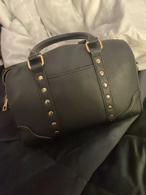 Svart handväska med nitar och gulddragkedja - Säljer en svart handväska i skinnimitation med guldfärgade detaljer och nitar på framsidan. Väskan har ett rejält handtag och dragkedja upptill. Perfekt storlek för det viktigaste och ger en cool vibe till din outfit.