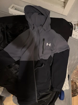 Under Armour vindjacka med huva - Snygg tvåfärgad vindjacka från Under Armour i mörkgrått och svart. Jackan har huva med snörning, bröstficka med dragkedja och logga på framsidan. Perfekt för blåsiga dagar och outdoor-aktiviteter. Materialet är polyester och jackan har normal passform.