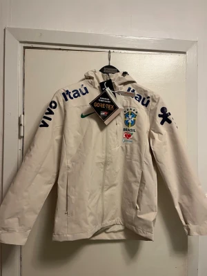 Beige Brazil Nike Gore-Tex vindjacka - Säljer en beige vindjacka från Nike med Brazil-logga och sponsormärken. Jackan är tillverkad i Gore-Tex-material, har huva och dragkedja framtill. Perfekt för dig som gillar fotboll och vill ha en sportig look. Snygga detaljer med tryck på ärmar och bröst.finns i storlek S-M-L 