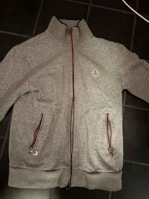 Grå Moncler zip hoodie - Säljer en grå hoodie från Moncler med hel dragkedja och ribbade muddar. Tröjan har Moncler-logga på bröstet och detaljer i rött, vitt och blått vid dragkedjorna. Perfekt för dig som gillar stilrena och exklusiva plagg.