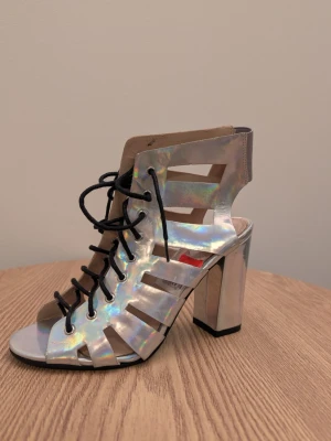 Silvriga holografiska klackade sandaler - Unika sandaler i holografiskt silver med bred klack och snörning framtill. Skon har öppen tå och coola utskurna detaljer på sidorna. Tillverkad i syntetmaterial och passar perfekt för dig som vill sticka ut. Finns ett litet skav märke på ena skon som du kan se på bilden