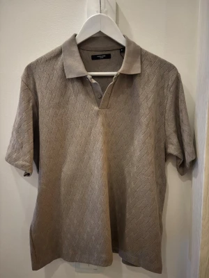 Beige stickad polotopp från Jack & Jones - Säljer en beige stickad polotopp från Jack & Jones i storlek M. Toppen har korta ärmar, krage och knappslå framtill. Materialet är stickat med diskret mönster som ger en snygg struktur. Perfekt till jeans eller chinos för en clean look.