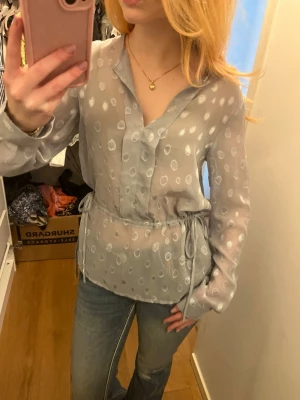Genomskinliga blå/grå blus  - Man kan välja hur indragen den ska vara i midjan och om man vill ha knappen knäppt i halsen. Den är ganska genomskinlig och de stora prickarna är minde genomskinliga.