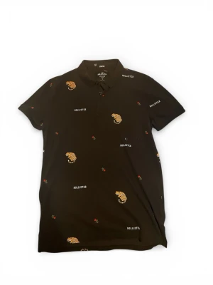 Svart Hollister piké med tigertryck - Svart pikétröja från Hollister med broderade tigrar, små röda rosor och logotypen utspridda över hela plagget. Klassisk krage och knappar framtill. Tillverkad i stretchigt bomullsmaterial för extra komfort.