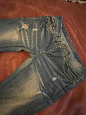 Blå bootcut jeans från ONLY med slitningar - Säljer ett par blå bootcut jeans från ONLY med slitna detaljer och dragkedjor på bakfickorna. Jeansen har en ljus tvätt med slitningar och hål på benen för en riktigt edgy look. Klassisk femficksmodell och bredare benslut. Perfekta för dig som gillar vintagevibbar.