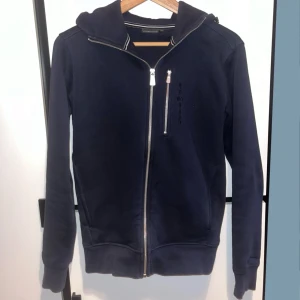 Marinblå hoodie från Sail Racing - Snygg marinblå hoodie från Sail Racing med dragkedja i silver och huva. Tröjan har en extra bröstficka med dragkedja och diskret logga på bröstet. Perfekt för dig som gillar sportig och stilren look. Storlek XS passar 170cm