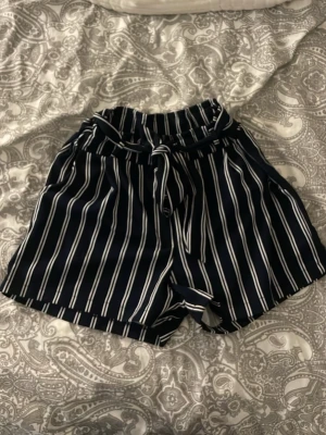 Randiga shorts från New Collection - Snygga högmidjade shorts i marinblått med vita vertikala ränder från New Collection. De har ett brett midjeband med knytband framtill och en loose passform. Perfekta för sommardagar och ger en chill men ändå stilren vibe. Storleken står inte på men är ganska stora fast passar ganska många olika eftersom att det är ett knytband. Jag är 167cm och de passar mig