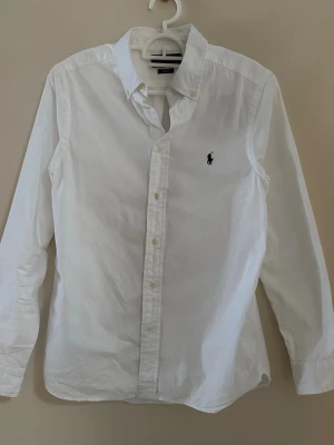 Vit skjorta från Polo Ralph Lauren - Vit skjorta från Polo Ralph Lauren i slim fit med klassisk krage och knappar framtill. Skjortan har lång ärm och den ikoniska broderade loggan på bröstet. Tillverkad i mjuk bomull som känns skön mot huden. Perfekt för dig som gillar stilrena och tidlösa plagg. Priset kan sänkas vid snabb förhandling!😄