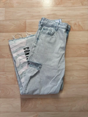 Ljusblå wide jeans med tryck och slitning - Unika ljusblå jeans från Bershka med bred passform och råa, fransiga benslut. Ena benet har stort tryck i svart och beige med texten 'CONTENT MIGHT BE DISTURBING BY SUNLIGHT'. Slitning vid fickan ger extra edge. Perfekta för dig som vill sticka ut.