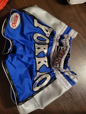 Blå och silver Yokkao Muay Thai shorts carbon  - Snygga Muay Thai shorts från Yokkao i blått och silver med stora broderade bokstäver och elastisk midja. Shortsen har svarta och vita detaljer längs kanterna och som är perfekt för träning.