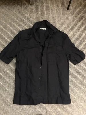Svart kortärmad skjorta från Zara - Svart skjorta från Zara i storlek S, tillverkad i linne. Skjortan har klassisk krage, korta ärmar och knäppning framtill med svarta knappar. Perfekt för dig som gillar enkel och stilren design.