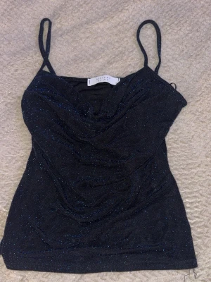 Svart/blå glittrig linnetopp Sisters Point - Säljer en svart/blå glittrig linnetopp från Sisters Point i storlek XS. Toppen har tunna axelband och ett snyggt draperat tyg framtill. Perfekt för dig som vill ha något extra till din outfit! Glittret är marinblått.