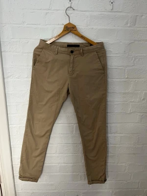 Beige chinos från Scotch & Soda - Snygga beige chinos från Scotch & Soda med klassisk passform och raka ben. Byxorna har bälteshällor, sidofickor och stängs med knapp och dragkedja. Tillverkade i mjuk bomull som känns skön hela dagen.