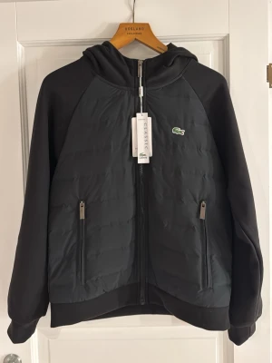 Svart pufferjacka från Lacoste - Säljer en svart pufferjacka från Lacoste med huva och dragkedja framtill. säljer för köpte två likadana med olika storlekat för se vilka som passar, detta blev den kvarblivna. fraktades en längre period av tid han ej returnera. Jackan har två fickor med dragkedja och den klassiska Lacoste-loggan på bröstet. Materialet är Bomull och jackan har en quiltad look som ger extra streetvibe. SKRIV VID MINSTA LILLA FRÅGA!!