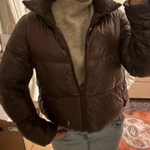 Pufferjacka - Säljer en brun pufferjacka med glansig finish och hög krage. Jackan har en rak, lite kortare passform och stängs med dragkedja framtill. Perfekt för kyliga dagar och enkel att styla med jeans eller hoodie.