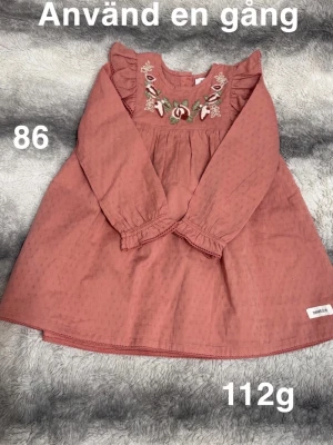 Rosa broderad klänning Newbie - Söt rosa klänning från Newbie med lång ärm och volangdetaljer vid axlarna. Broderade blommor framtill och diskret prickigt mönster i tyget. Klänningen har rund hals. perfekt för vår och sommar.🌸 Använd en gång. Storlek 86