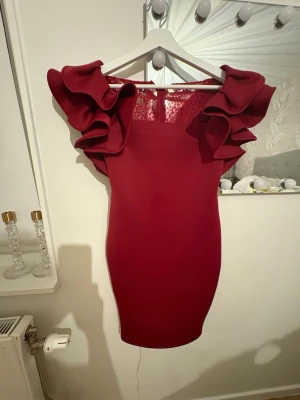 Vinröd kortklänning med volangärmar - Supersnygg vinröd kortklänning med stora volangärmar och spetsdetaljer upptill. Klänningen har en tight passform och dragkedja i ryggen. Perfekt för dig som vill sticka ut med en unik look och älskar statement-ärmar.