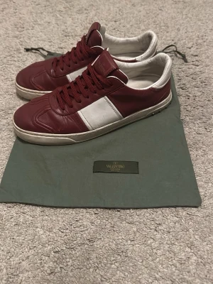 Valentino Flycrewr - Säljer ett par snygga Valentino Flycrew sneakers i vinrött och vitt skinn. Skorna har klassisk snörning, rund tå och vita detaljer på sidan och hälen. Sulan är vit med nitar baktill och svart mönster undertill. Perfekta för dig som vill ha något exklusivt och stilrent. Två nitar saknas. Helt okej skick. Kanske byten