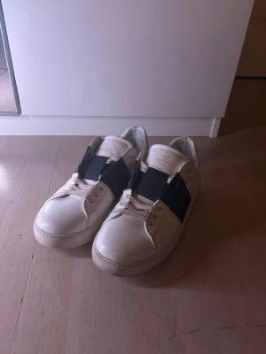 Valentino Garavani vita sneakers - Säljer ett par vita Valentino Garavani sneakers i skinn med bred svart rem över mitten och klassisk snörning. Skorna har rund tå och platt sula, och kommer med originalpåse och extra skosnören. Perfekta för dig som gillar stilrena och exklusiva sneakers. Skorna kommer även med en golden goose påse som inte används!!!