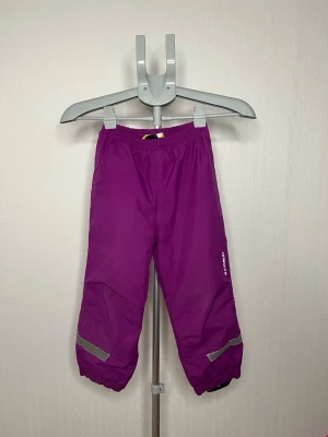 Lila täckbyxor från Everest 110/116 - Säljer ett par lila täckbyxor från Everest i storlek 110/116. Byxorna har resår i midjan och benslut, samt reflexdetaljer vid benen för extra synlighet. .