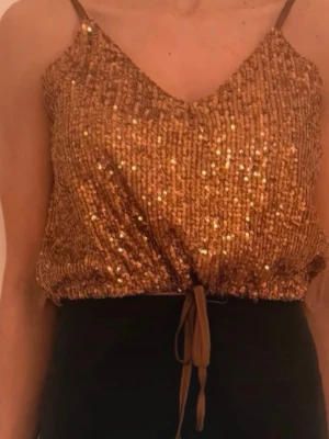 Glittrig guld tubtopp med snörning - Säljer en glittrig guld tubtopp med smala axelband och snygg snörning framtill. Toppen har en croppad passform och är täckt av glittriga paljetter som verkligen sticker ut. Perfekt för dig som vill ha något extra till kvällen!