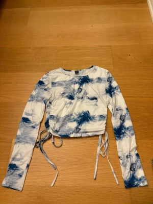 Blåvit croppad topp från SHEIN, storlek XS - Säljer en croppad långärmad topp från SHEIN i storlek XS. Toppen har ett blåvitt abstrakt mönster och snörning i sidorna för justerbar passform. Materialet är mjukt och stretchigt, perfekt till jeans eller kjol. 100% Polyester.