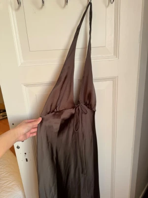 Brun halterneckklänning från Zara - Snygg brun långklänning från Zara i satinliknande material. Klänningen har halterneck, knytning framtill och en elegant, slät yta som ger en lyxig känsla. Ny med prislapp utan defekter. 