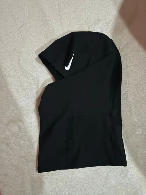 Svart Nike Pro balaclava - Säljer en svart Nike Pro balaclava med vit swoosh-logga på sidan. Tillverkad i stretchigt syntetmaterial som andas, perfekt för träning utomhus. Sitter tight runt huvudet och har en clean, sportig look.