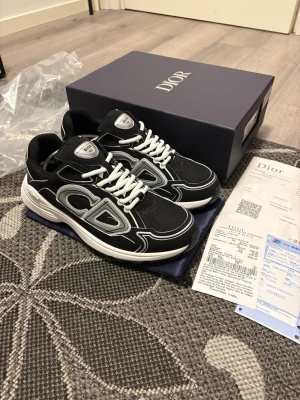 Svarta Dior sneakers med vita detaljer - Säljer ett par svarta Dior sneakers med vita och silverfärgade detaljer samt stora CD-loggor på sidorna och hälen. Skorna har vit platt sula, snörning och meshpaneler för extra andningsförmåga. Riktigt snygga och trendiga sneakers som sticker ut.