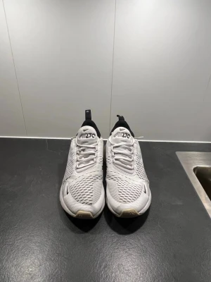 Nike 270 - Nike Air Max 270 sneakers i vit färg med svarta detaljer och synlig luftkudde i hälen. Skorna har mesh-överdel för bra ventilation, snörning och rund tå. Perfekta för dig som gillar sportig stil och vill ha bekväma sneakers med snygg design.