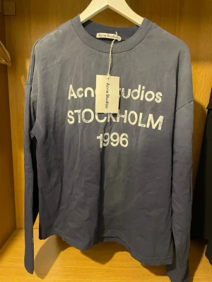 Blå långärmad t-shirt Acne Studios - Snygg blå långärmad t-shirt från Acne Studios med tryck 'Acne Studios Stockholm 1996' i vitt framtill. Tillverkad i 100% bomull och har en avslappnad passform. Perfekt för dig som gillar streetwear och skandinavisk design.