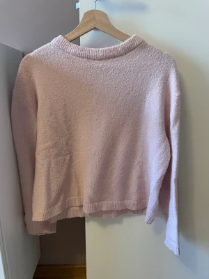 Ljusrosa stickad tröja - Mysig ljusrosa stickad tröja med rund halsringning och långa ärmar. Tröjan har en enkel, avslappnad passform och ribbade muddar vid hals, ärmslut och nederkant. Perfekt för lager på lager under kyliga dagar.