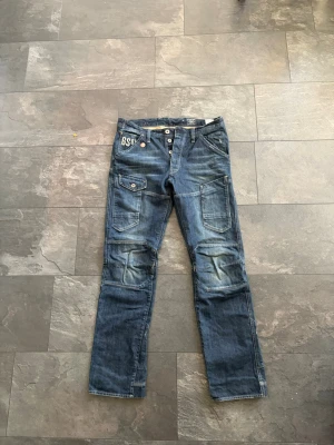 G-Star RAW blå cargopants jeans - Säljer ett par blå cargopants från G-Star RAW med flera stora fickor och coola detaljer. Hittad i farsans garderob. Jeansen har knappgylf, kontrastsömmar och broderad logga vid fickan. Materialet är slitstarkt jeans och passformen är loose med raka ben. Helt nya, bortsett från att de köptes för ett par år sen.