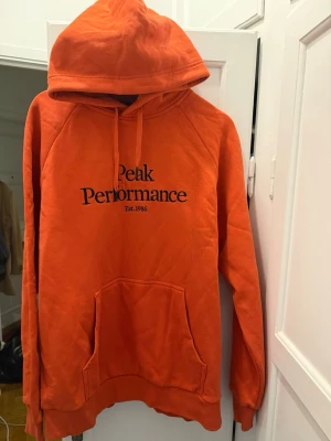 Peak Performance Orange Hoodie - En tjock skön hoodie med bra kvalité som är perfekt nu vid vinter tider! Hoodien har 52cm axelbredd och 64 cm i längd. 