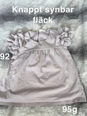 Ljuslila blus med volangärmar - Supersöt ljuslila blus från POMPdeLUX med stora volangdetaljer på axlarna. Blusen har rund halsringning och knapp i nacken. Perfekt för dig som gillar romantiska detaljer och mjuka färger.🌸 hittade en knappt synbar fläck när jag synade inför försäljning. Storlek 92