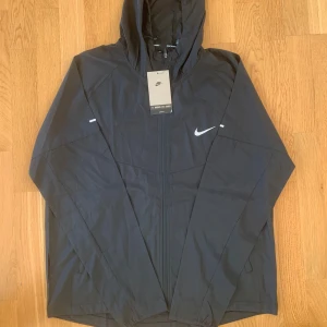  Nike windbreaker - Svart windbreaker från Nike med luva. Jackan har dragkedja framtill och är tillverkad i lätt polyester, perfekt för löpning eller andra utomhusaktiviteter. Snygg och sportig design. Pris kan diskuteras.