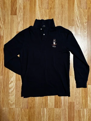 Mörkblå  långärmad Polo Ralph  - Mörkblå långärmad skjorta från Polo Ralph Lauren i custom slim fit. Skjortan har broderad björn med POLO-text på bröstet, klassisk krage och två knappar vid halsen. Tillverkad i mjuk bomull som är skön mot huden. Perfekt för dig som gillar stilrena och ikoniska plagg.