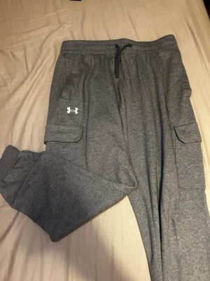 Grå mjukisbyxor från Under Armour - Säljer ett par grå mjukisbyxor från Under Armour med bekväm resårmidja och snörning. Byxorna har praktiska cargofickor på sidorna och ribbade muddar vid bensluten. Perfekta för chill eller träning, med stilren logga på benet.