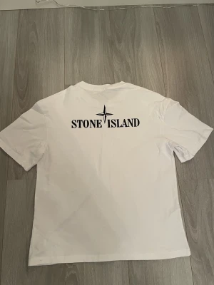 Vit Stone Island t-shirt med logga - Vit t-shirt från Stone Island i bomull med klassisk rund hals och svart logotyptryck på bröstet samt stort tryck på ryggen. Snygg och enkel design som passar perfekt till jeans eller shorts. Märkesetikett i nacken och autenticitetslapp på insidan.