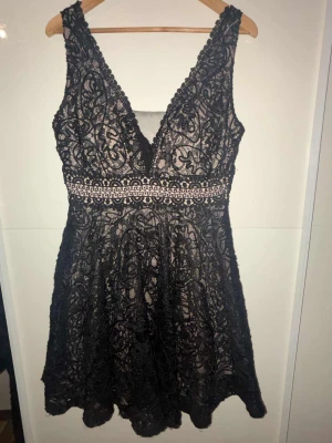 Svart spetsklänning med v-ringning - Snygg kort svart klänning med djup v-ringning och spetsdetaljer över hela klänningen. Midjan har ett dekorativt band med mönster och klänningen är ärmlös. Perfekt för dig som gillar detaljer och vill sticka ut med en elegant look.