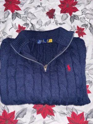 Marinblå stickad tröja Polo Ralph Lauren - Säljer en marinblå stickad tröja från Polo Ralph Lauren med halv dragkedja och klassiskt rött broderat logomärke på bröstet. Tröjan har kabelstickat mönster, ribbade muddar och lång ärm. Perfekt för kyliga dagar och snygg till jeans.