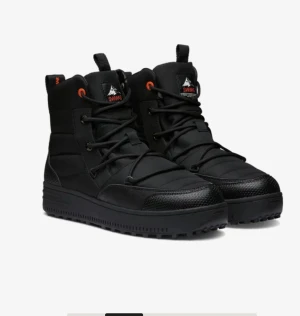Svarta vinterboots från Swims - Säljer ett par svarta boots från Swims med snörning och orange detaljer vid öglorna. Skorna har en robust platt sula och quiltad ovandel i syntetmaterial, perfekt för kalla dagar. Märkeslogga på plösen och dragflik baktill för enkel påtagning.