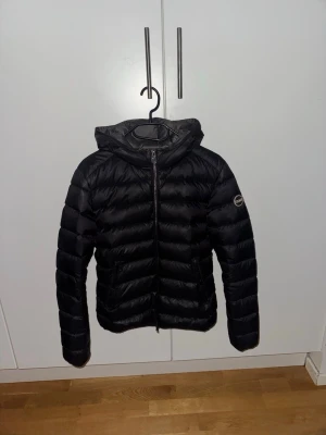 Svart Colmar dunjacka med huva - Svart dunjacka från Colmar med huva och dragkedja framtill. Jackan har ett quiltat mönster och två sidofickor. Perfekt för kyliga dagar och har en stilren look med Colmar-logga på ärmen. Materialet är syntet och dun, vilket gör den både lätt och varm.