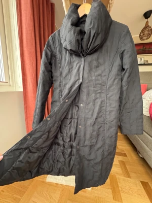 Svart lång insvängd dunjacka - Säljer en svart lång dunjacka med quiltad struktur och stor, mysig sjalkrage. Jackan har knappar framtill och insvängda sömmar. Väldigt lätt att ta av och på! Lite tunnare design jämfört med pufferjackor.